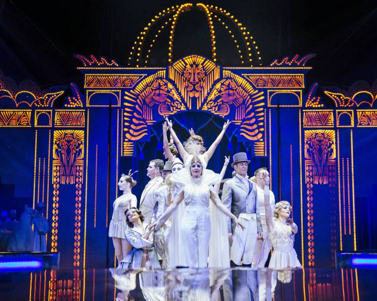 Viele Schauspieler in weißer Kleidung vor einer blau-orangenen Bühnenkonstruktion im Circus Krone.