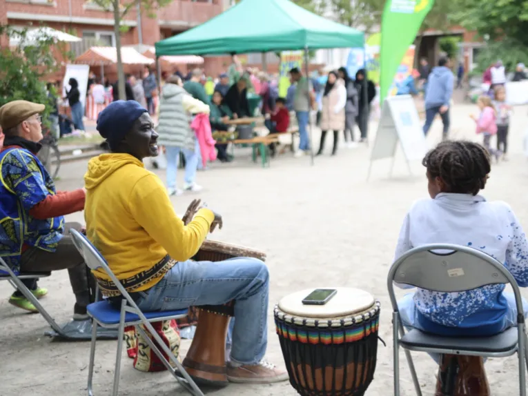 Zwei M&auml;nner mit Trommeln sitzen auf St&uuml;hlen. Vor ihnen ein Platz mit Menschen, rechts neben ihnen ein Kind auf einem Stuhl.