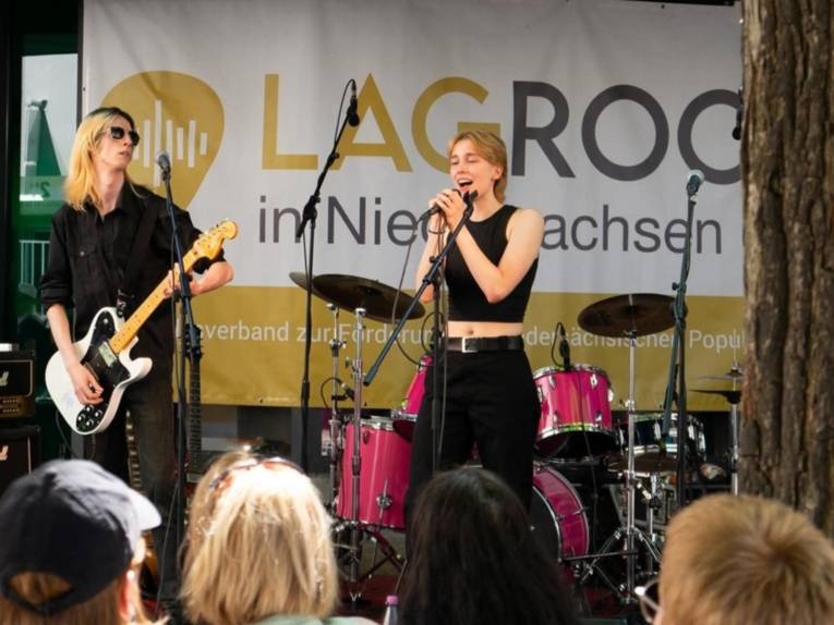 Mann mit Gitarre steht auf einer Bühne. Neben ihm eine Frau mit Mikrofon. Beide stehen vor einem Publikum.