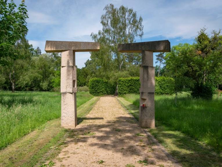 Eine Art steinernes Portal auf einem Friedhof. Davor stehen Kerzen.