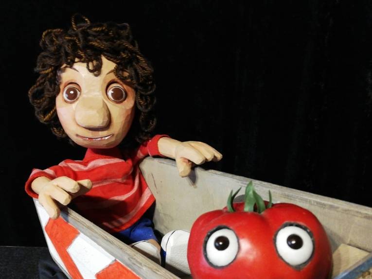 Zu sehen ist eine Marionette, die in einem Karren sitzt. Davor befindet sich eine große Tomate.