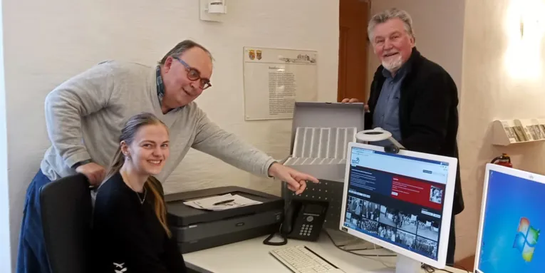 Ein Mann und eine Frau schauen auf einen PC-Bildschirm, hinter dem Bildschirm steht ein weiterer Mann und schaut in die Kamera.