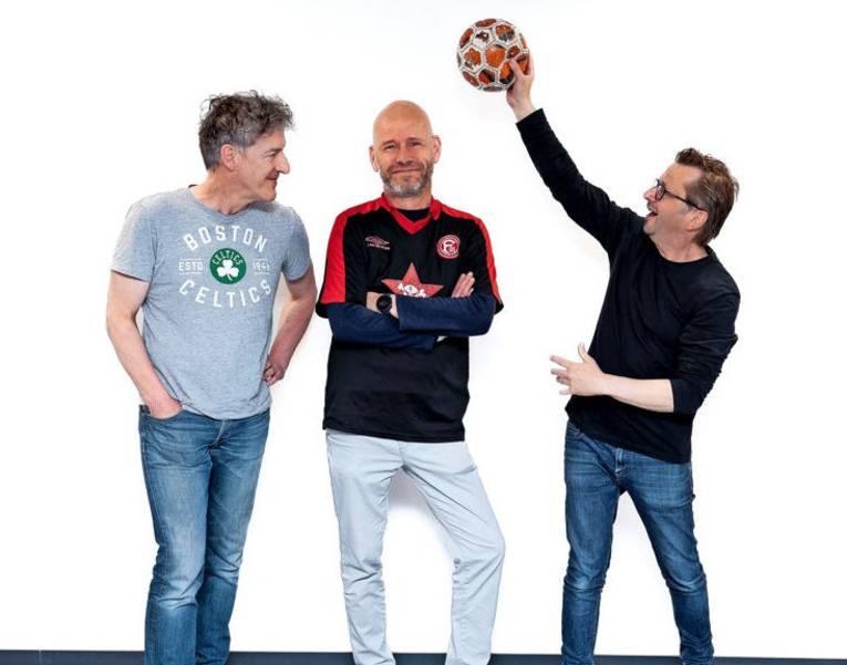 Drei Männer, einer hält einen Ball in die Höhe.