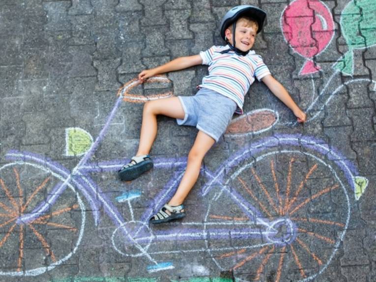 Kreidezeichnung eines Fahrrads auf der Straße, ein Junge mit Helm, der darauf liegt, als würde er auf dem Sattel sitzen