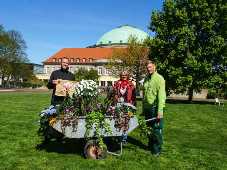 Drei Personen hinter einer Schubkarre voll Blumen auf einer grünen Wiese. Im Hintergrund das hannoversche HCC.