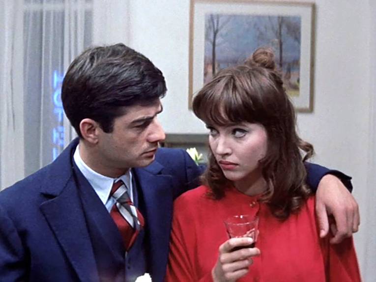 Ein Mann in Anzug und Krawatte legt seinen Arm um eine Frau. Sie hat ein kleines Glas in der Hand und schaut ihn skeptisch an.