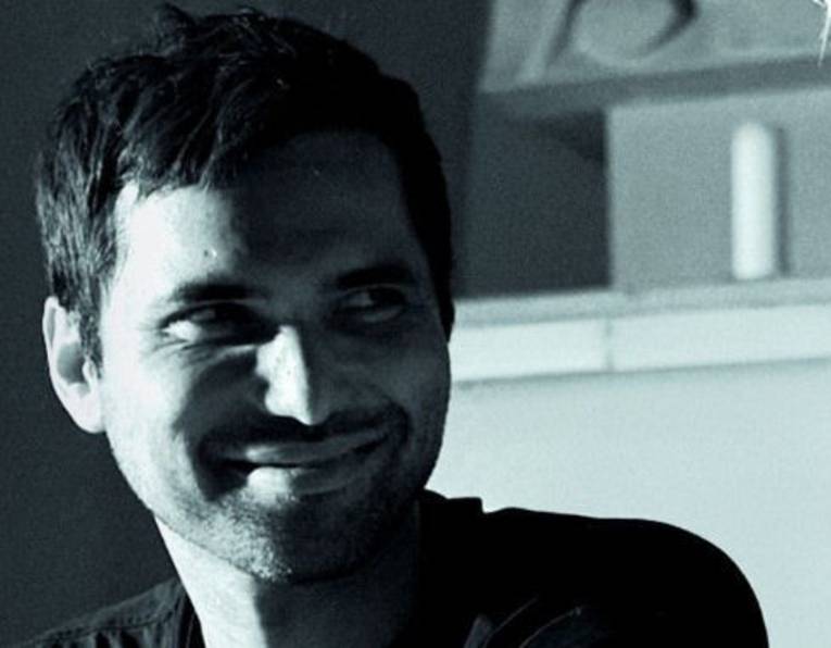 Nahporträt eines lächelnden Mannes mit kurzen Haaren und Bart, seine linke Gesichtshälfte ist verschattet.