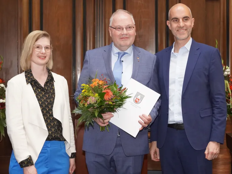 Drei Personen stehen frontal zur Kamera in einem Raum: In der Mitte ein Mann mit einem Blumenstrauß und einer Urkunde, links eine Frau, rechts ein Mann.