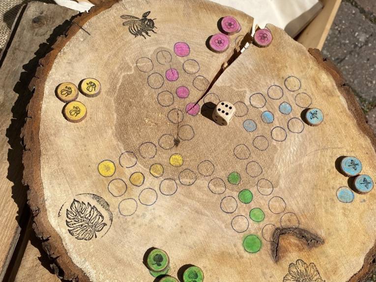 Auf eine Baumscheibe ist das Spielbrett eines Würfelspiels gezeichnet. Felder sind farbig markiert und Stempel verzieren das Spielbrett.
