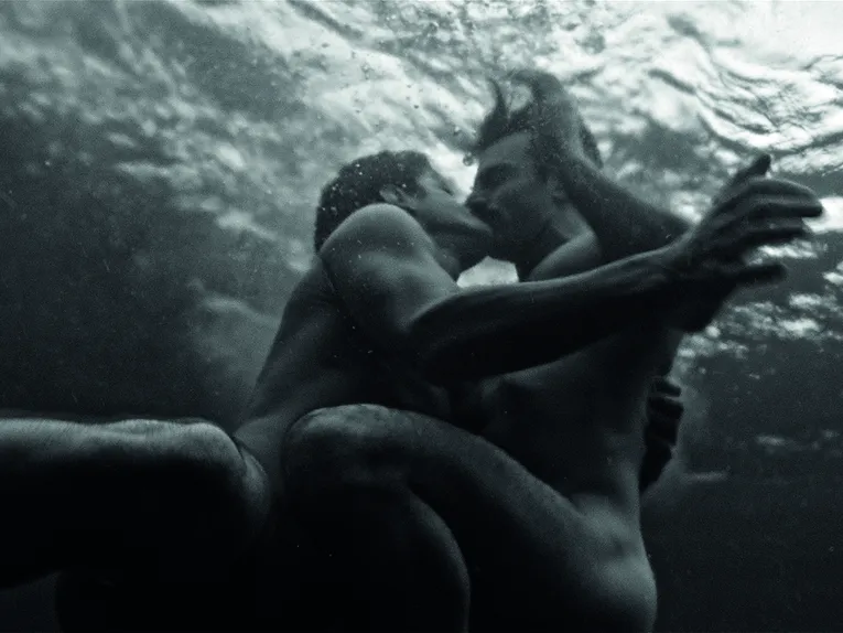 Das Schwarz-Weiß-Bild zeigt zwei Personen unter Wasser in einer innigen Umarmung. Die Aufnahme wurde aus einer Perspektive unterhalb der Wasseroberfläche fotografiert, wodurch Lichtreflexe und die Bewegung des Wassers eine traumartige Atmosphäre erzeugen. Die Körper der beiden Personen sind nur teilweise sichtbar und verschmelzen mit den weichen Kontrasten der Umgebung zu einer poetischen, künstlerischen Komposition.