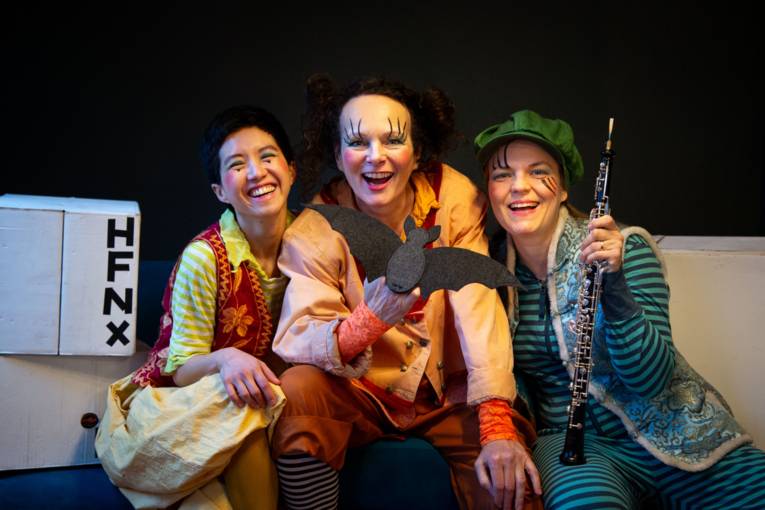 Drei Menschen in bunten Kostümen sitzen nebeneinander und lächeln in die Kamera.