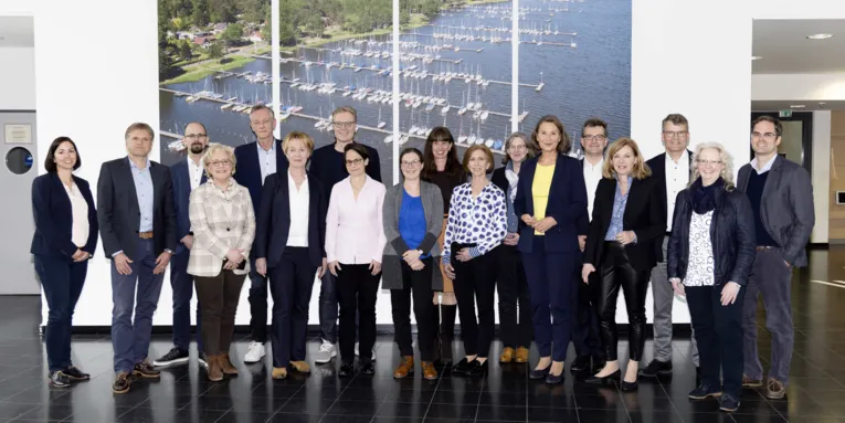 Gruppe von Personen vor einem Bild auf dem ein Gewässer, Bootsanleger und Segelboote zu sehen sind.