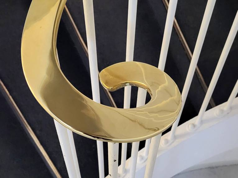 Ende eines weißen Treppengeländers an einer schwarzen Treppe, der Griff in Gold dreht sich ein wie ein Schnörkel.