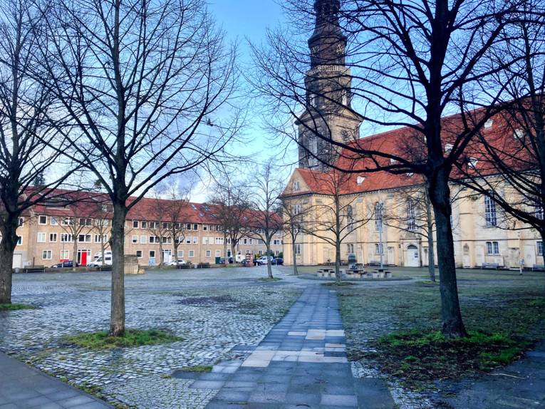 Ein gepflasterter Platz mit vier kahlen Bäumen, links hinten eine Häuserreihe, rechts eine Kirche, der Kirchturm mittig hinten im Bild.