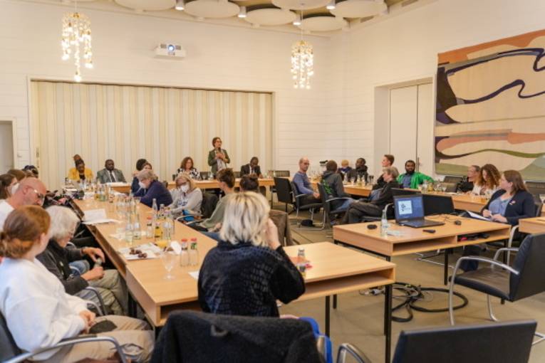 Mehrere Personen sitzen in einem Raum an Tischen und h&ouml;ren einer stehenden Person zu.