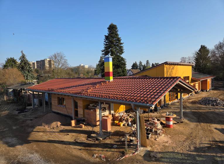 Ein Gebäude mit einem Geschoss, roten Ziegeln und einem farbig gestreiften Schornstein von seitlich oben fotografiert. Drum herum ein sandiger Weg, Steinhaufen und Baumaterialien wie Bretter udn Eimer.