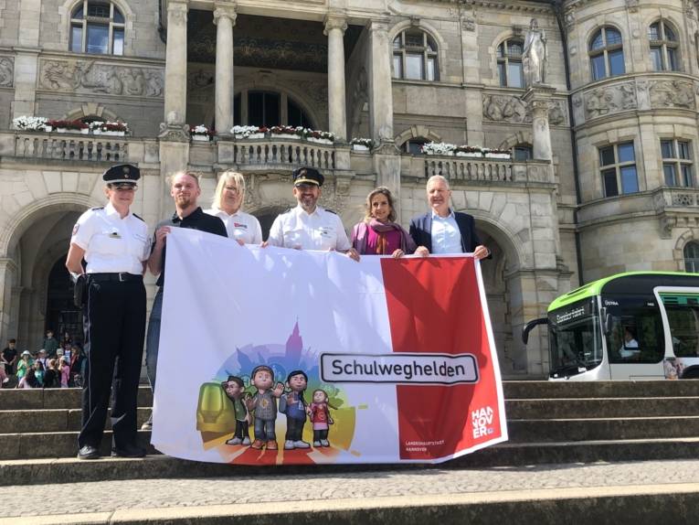 Sechs Personen stehen vor dem Neuen Rathaus und halten ein Plakat mit der Aufschrift "Schulweghelden" in die Kamera.