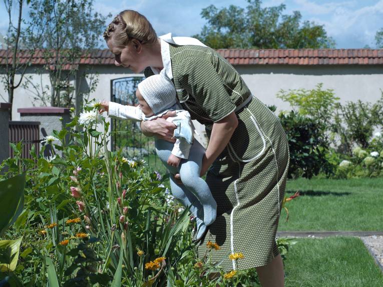 Das Bild zeigt eine Person, die ein Kleinkind in einem blühenden Garten vor einem Blumenbeet hält. Das Kind streckt die Hand nach einer weißen Blüte aus, während die Person sich zu den Pflanzen hinunterbeugt. Im Hintergrund sind eine Mauer, Bäume und gepflegte Grünflächen zu sehen. Das helle Tageslicht und die vielfältigen Farben der Blumen verleihen der Szene eine natürliche und ruhige Atmosphäre.