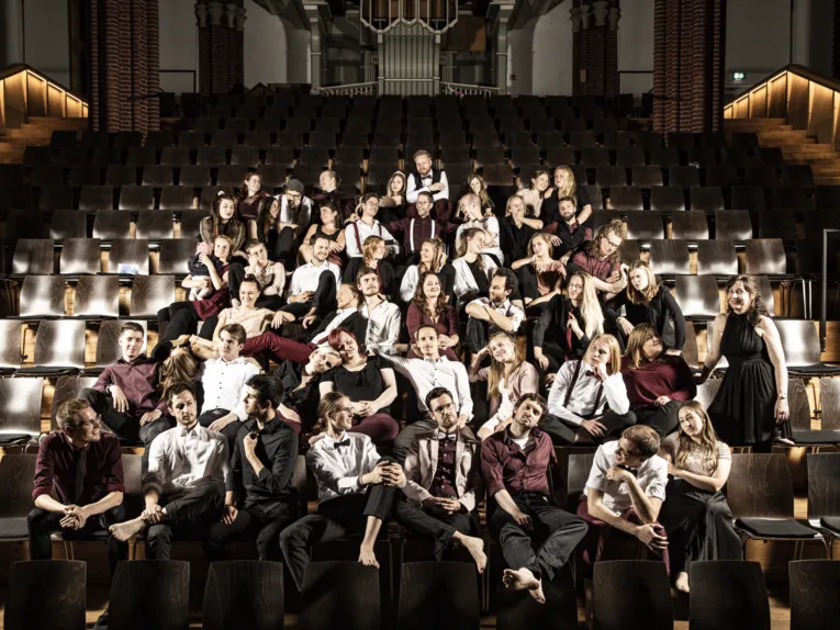 Der Vivid-Voices-Chor sitzt in den Sitzreihen eines Aufführungsortes.