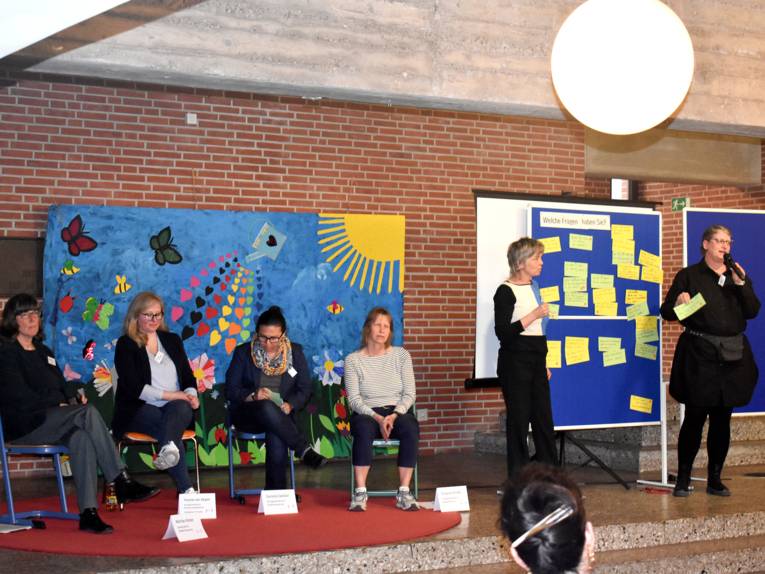 Vier Frauen sitzen auf St&uuml;hlen auf einem Podium, zwei Frauen stehen daneben an einer Tafel mit Zetteln.