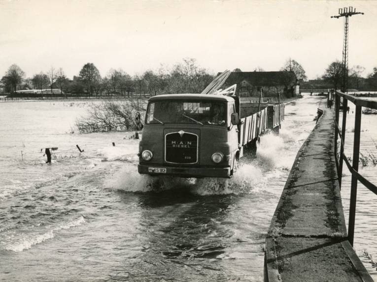 Historisches Schwarz-Weiß-Foto: Ein Lkw mit Anhänger fährt durch Wasser, überflutete Stacheldraht-Zäune und Bäume sind im Bild.