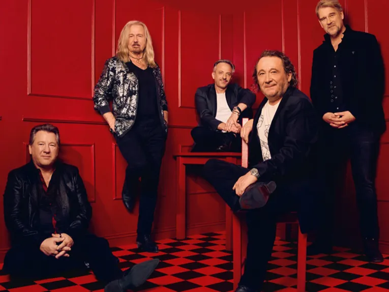 Zu sehen ist die deutsche Band Karat mit seinen fünf Mitgliedern in schwarzer Ausgehleidung in einem roten Raum.