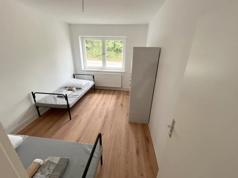 Blick in einen schmalen Raum, vorn links ein Bett, hinten links ein Bett, in der Mitte ein Fenster, rechts Spinde und rechts vorn eine offene T&uuml;r.