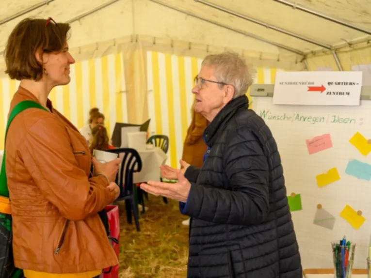 Eine Mittelalte und eine &auml;ltere Frau unterhalten sich in einem gelb-wei&szlig;en Zelt. Hinter Ihnen ein Flipchart mit dem Text W&uuml;nsche, Anregungen, Ideen. Weiter hinten sitzen zwei weitere Personen an einem Tisch.