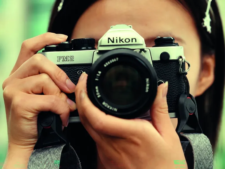 Das Bild zeigt eine Person, die eine analoge Spiegelreflexkamera vom Typ Nikon FM2 vor das Gesicht hält und durch den Sucher fotografiert. Die Kamera steht im Mittelpunkt der Aufnahme und wird von beiden Händen umfasst. Durch die Nahaufnahme sind Details des Kameragehäuses und des Objektivs deutlich sichtbar, während der Hintergrund unscharf bleibt. Die warmen Farben und die geringe Tiefenschärfe verleihen dem Bild eine konzentrierte, fotografische Atmosphäre.
