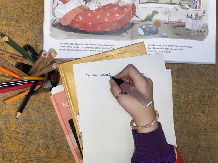 Zu sehen ist ein aufgeschlagenes Buch und ein Stapel Kinderbücher. Ebenfalls ist eine Hand zu sehen, die auf einem Blatt Papier: "Es war einmal..." schreibt.