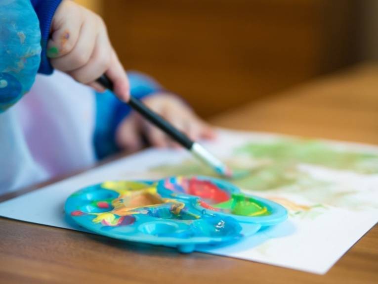 Bild einer Kinderhand mit Pinsel und Farbpalette, die ein Bild malt.