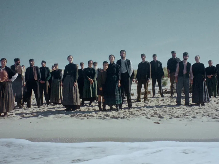 Eine Gruppe von Menschen in historisch wirkender, schlichter Kleidung steht am Strand in einer lockeren Reihe und blickt in Richtung Meer. Im Vordergrund sind Sand und die anrollenden Wellen zu sehen, während sich im Hintergrund ein weiter, wolkenloser Himmel über der Küstenlandschaft erstreckt. Die gedämpften Farben und die ruhige Komposition verleihen der Szene eine zeitlose, beinahe melancholische Atmosphäre.