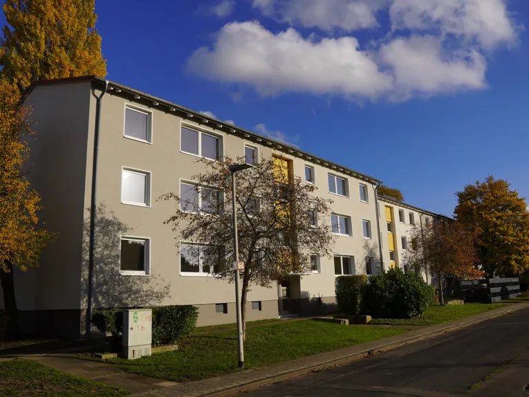 Ein langes Wohngebäude mit zwei Obergeschossen, davor Rasen und ein Baum, blauer Himmel mit weißen Wolken.