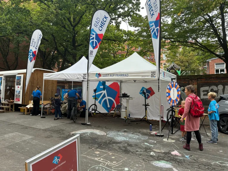 Ein Zelt mit der Aufschrift Lust auf Fahrrad, davor Beachflags. Unter dem Zeltdach stehen Erwachsene und Kinder &uuml;ber Fahrr&auml;der gebeugt.