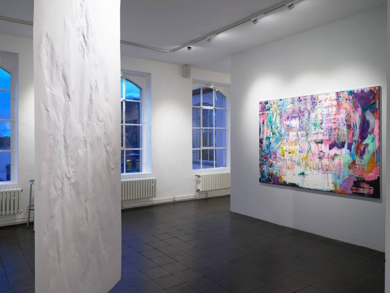 Zu sehen ist eine Raumsicht der Galerie Drees: Ein sehr buntes Bild hängt an einer weißen Wand, eine weiße Fläche hängt von der Decke.