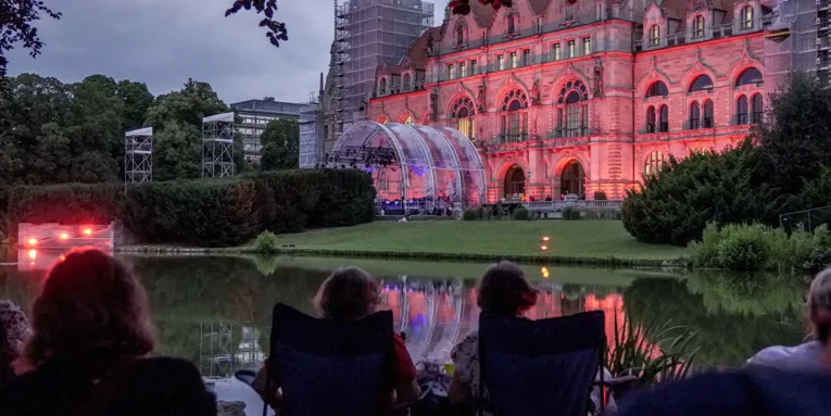Menschen in einem Park. Im Hintergrund eine beleuchtete Bühne.