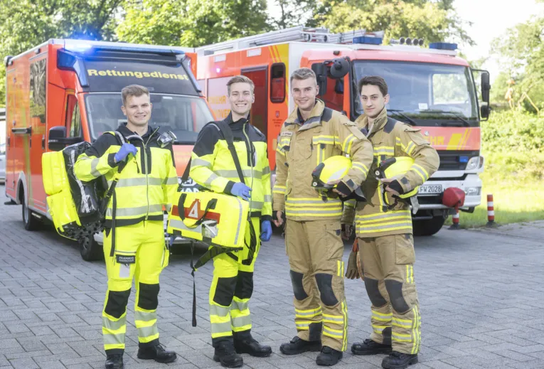 Vier Personen vom Rettungsdienst stehen in Arbeitskleidung lächelnd vor einem Rettungswagen und einem Feuerwehrwagen.