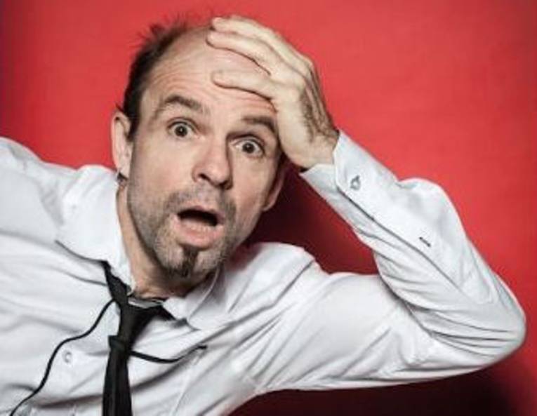 Mann mit kurzem Resthaar, Dreitagebart, hochgezogenen Augenbrauen und geöffnetem Mund hält sich die linke Hand an den Kopf.