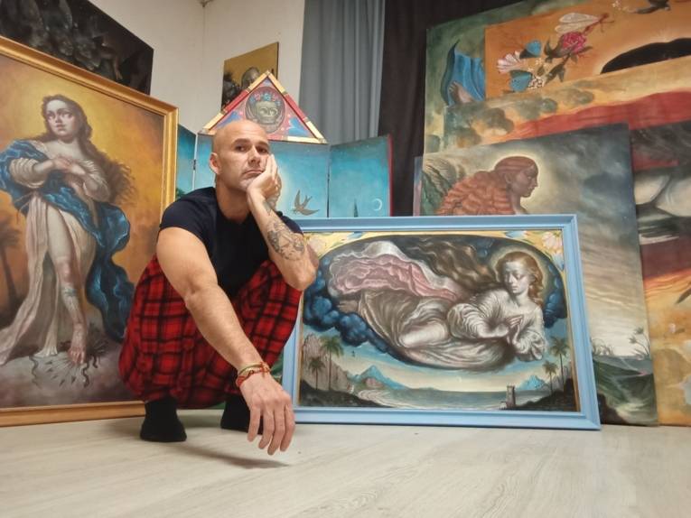 Ein Mann hockt auf dem Boden vor gemalten Madonnen-Bildern. Er stützt seinen Kopf auf eine Hand und schaut in die Kamera.