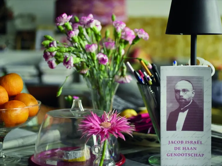 Ein liebevoll arrangiertes Stillleben zeigt einen Tisch mit Blumen, einer Schale Orangen, Schreibutensilien und einer Tischlampe. Im Vordergrund fällt ein Buch mit einem historischen Porträt auf, während die unscharfe Umgebung und das weiche Licht eine ruhige, persönliche Atmosphäre schaffen.