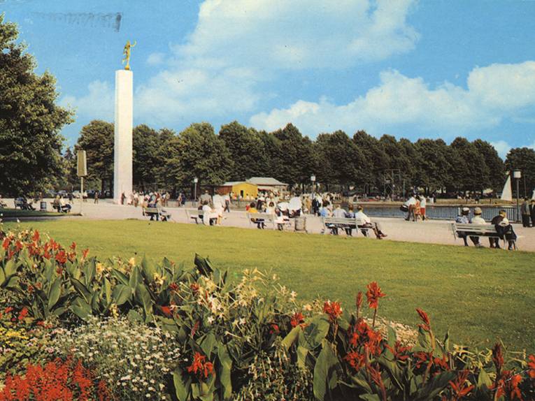 Farbige Foto-Postkarte, Poststempel 1974: Arthur-Menge-Ufer Nordufer des Maschsees . Im Vordergrund Blumenbeete, Blick auf Rasenfläche, auf Bänken sitzende Personen und auf die Fackelträger-Säule am Ufer des Sees. Rückseitig bezeichnet "HANNOVER Partie am Maschsee"