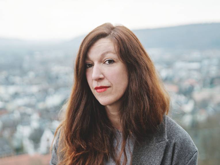 Eine Frau mit langen, rötlichen Haaren sitzt vor einem unscharfen Stadthintergrund.