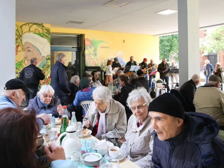 Vorne sitzt eine Gruppe &auml;lterer Menschen an einem Kaffeetisch und unterh&auml;lt sich. In der Mitte sitzt eine Gruppe mittelalter Menschen an einem Tisch. Im Hintergrund stehen sechs Menschen mit Instrumenten und einem Notenst&auml;nder vor sich.