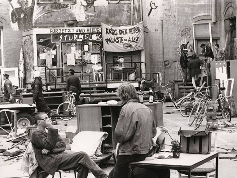 Gelände der Sprengel-Schokoladenfabrik, Foto von Eberhard Franke, um 1988 
 Hannover-Nordstadt, Schaufelderstraße. Entwicklung nach Krawallen und Hausbesetzungen zu einem alternativen Viertel. Aufnahme mit Blick Richtung Süden. Entstanden während der Besetzung der leerstehenden Gebäude an der Schaufelderstraße in den 1980er Jahren. Zu sehen sind Teile des Fabrikhofs und der Eingang zu den Gebäuden. Auf dem Hof stehen verschiedene Möbel, wie Aktenschränke und Spinde (vermutlich aus der Fabrik) sowie mehrere Fahrräder und Baumaterial/Bauschrott. Im Vordergrund drei Personen, vermutlich Mitglieder der Besetzer im Gespräch an einem Tisch. Im Hintergrund link sieht man drei Polizeibeamte in Uniform, einer der Beamten mit Schutzausrüstung, die das Gebäude begutachten. Rechts im Bild auf einer Treppe eine weitere Gruppe mit Polizisten. Im Zentrum der Aufnahme der verbarikadierte Eingang mit einem daneben platzierten Transparent: "Krieg den Hütten Paläste für alle", NANA-Archiv im HMH