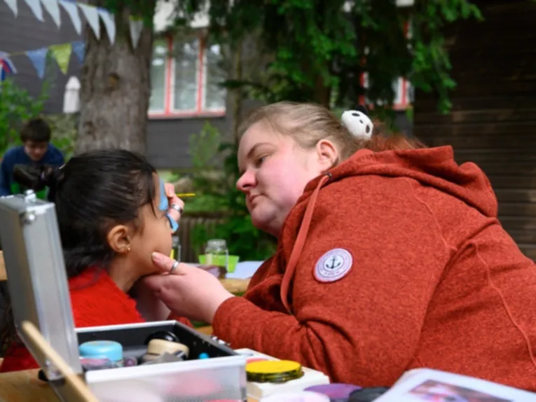 Eine Frau bemalt das Gesicht eines M&auml;dchens. Vor ihnen ein kleiner Koffer mit Farben.
