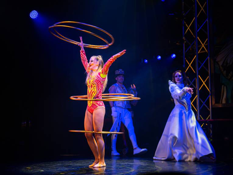 Eine Artistin lässt auf einer Bühne mehrere Hula-Hoop-Ringe um ihren Körper kreisen.