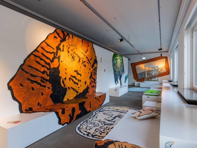Blick in das Mitmachfoyer SammleR*s: Die weißen Wände sind zum Teil bedeckt mit großflächigen bunten Paste-ups von Objekten aus dem Museum. So auch die Kissen, die auf der weißen Sitzaldnschaft liegen. Am Ende des schmalen Ganges im Bildhintergrund sieht man eine Vitrine mit kleinen Figuren.