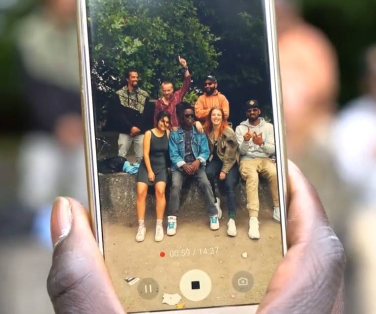 Eine Hand hält ein Smartphone, dessen Kamera aktiv ist. Auf dem Bild der Kamera sind sieben Menschen zu sehen, vier sitzen auf einer niedrigen Mauer, drei stehen dahinter.