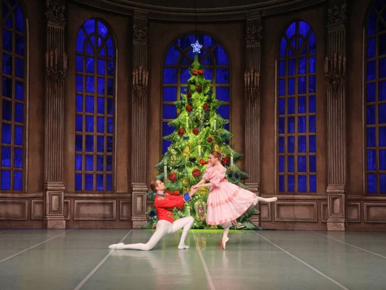 Zu sehen ist eine Bühnenszene aus der "Nussknacker": Ein großer Weihnachtsbaum im Hintergrund, ist im Vordergrund ein tanzendes Paar zu sehen.