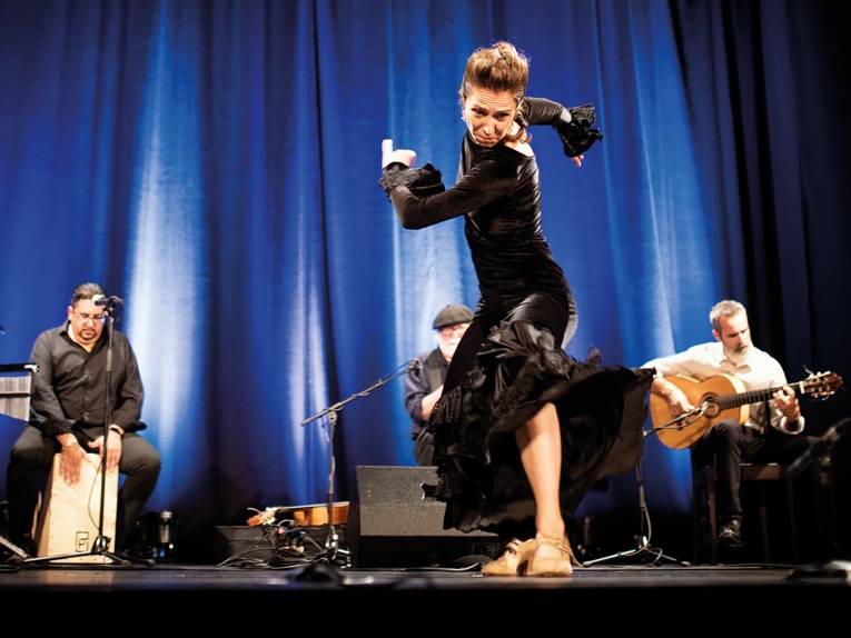 Eine Frau im schwarzen Kleid tanzt Flamenco, im Hintergrund sieht man Musiker*innen.